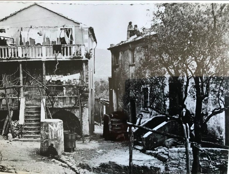 Fotografia d’epoca in bianco e nero della piazzetta di Reggimonti: due case in pietra con il bucato steso, una vasca in pietra, un albero e una persona seduta sugli scalini.