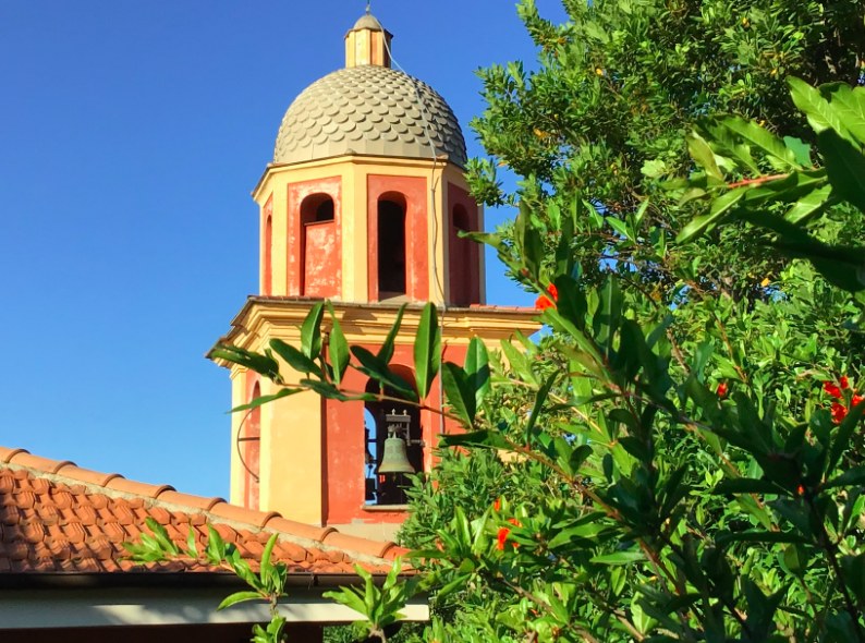 Il campanile della chiesa con la cupola rivestita di scaglie grigie, i decori ocra e terracotta e la campana in bronzo visibile nell'arcata; in primo piano i coppi di un tetto e i rami di un melograno sotto un cielo sereno.